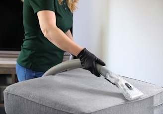 a woman in a green shirt and black gloves vacuuming a gray ottoman