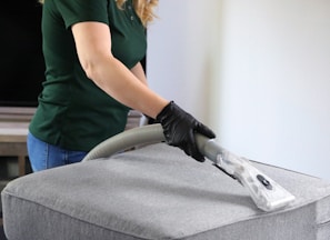 a woman in a green shirt and black gloves vacuuming a gray ottoman