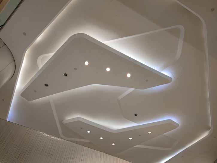 A modern ceiling design featuring sleek, geometric shapes with embedded lights. The ceiling has a minimalistic and futuristic appearance with smooth, overlapping panels that create depth.