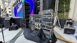 A stage setup with various electronic equipment, including amplifiers and cables, organized on racks. There are large speakers on stands, a big screen displaying colorful graphics, and wrapped cables. The stage area is covered in black cloth. Some green plants can be seen on the right-hand side, and the scene appears to be in an open or semi-open area with a mix of artificial and natural lighting.