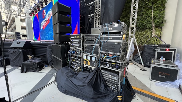A stage setup with various electronic equipment, including amplifiers and cables, organized on racks. There are large speakers on stands, a big screen displaying colorful graphics, and wrapped cables. The stage area is covered in black cloth. Some green plants can be seen on the right-hand side, and the scene appears to be in an open or semi-open area with a mix of artificial and natural lighting.