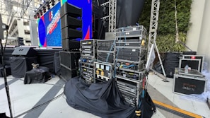 A stage setup with various electronic equipment, including amplifiers and cables, organized on racks. There are large speakers on stands, a big screen displaying colorful graphics, and wrapped cables. The stage area is covered in black cloth. Some green plants can be seen on the right-hand side, and the scene appears to be in an open or semi-open area with a mix of artificial and natural lighting.