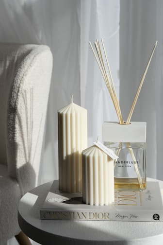 A cozy setup featuring two cream-colored, fluted candles placed on a round side table. Adjacent to the candles is a diffuser bottle with wooden reeds, labeled with 'Wanderlust New York.' Underneath these items is a Christian Dior fashion book, all placed in a softly lit room with a white curtain background.