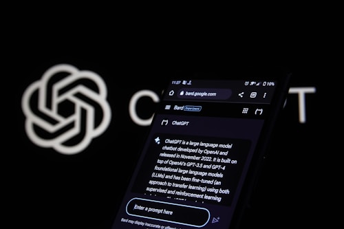 A smartphone screen displays a web page about ChatGPT, featuring a black background with white text. The screen includes a search bar and various interface icons at the top. In the background, an OpenAI logo is partially visible against a black backdrop.