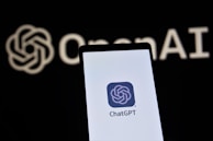 A smartphone screen displaying the ChatGPT logo in focus, with the OpenAI text and logo faintly visible in the background.