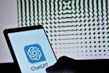A smartphone displaying the ChatGPT logo is held against a backdrop of a geometric pattern, which consists of wavy, radial green lines that create an illusion of depth.