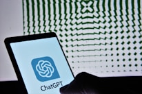 A smartphone displaying the ChatGPT logo is held against a backdrop of a geometric pattern, which consists of wavy, radial green lines that create an illusion of depth.