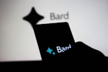 A smartphone held by a person displays the word 'Bard' with a stylized star shape logo above it, while in the background, a similar logo and the word 'Bard' appear on a large screen.