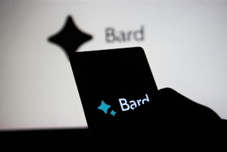 A smartphone held by a person displays the word 'Bard' with a stylized star shape logo above it, while in the background, a similar logo and the word 'Bard' appear on a large screen.