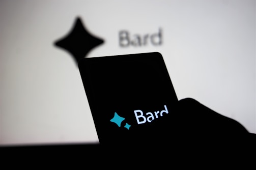 A smartphone held by a person displays the word 'Bard' with a stylized star shape logo above it, while in the background, a similar logo and the word 'Bard' appear on a large screen.
