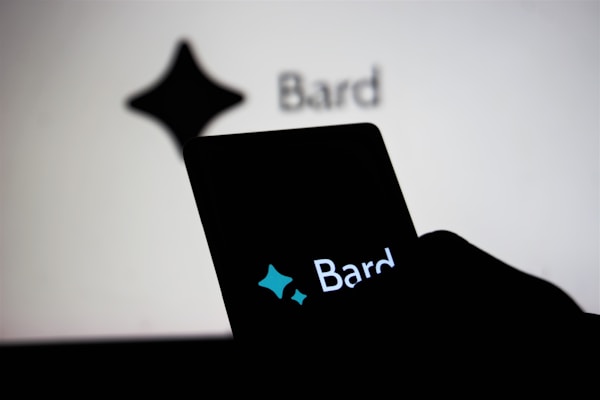 A smartphone held by a person displays the word 'Bard' with a stylized star shape logo above it, while in the background, a similar logo and the word 'Bard' appear on a large screen.