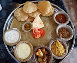 A traditional Indian thali is arranged with various dishes served on a large metal plate. The plate contains several small bowls filled with different curries, a dessert, pickles, and rice. There are also a few pieces of puffed bread known as puris and some sliced tomatoes placed in the center.