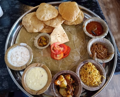 A traditional Indian thali is arranged with various dishes served on a large metal plate. The plate contains several small bowls filled with different curries, a dessert, pickles, and rice. There are also a few pieces of puffed bread known as puris and some sliced tomatoes placed in the center.