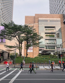 A bustling urban scene with several people walking across a crosswalk in front of tall modern buildings. The lush greenery of trees and hedges adds a touch of nature to the city environment. Pedestrians include individuals casually dressed, some in activewear and others in leisure clothing. The buildings are characterized by large windows and a sleek, contemporary design.