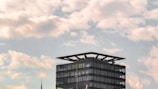 A modern glass-clad building with a distinctive flat roof structure set against a backdrop of a partly cloudy sky. Below the clouds, the building's simple geometric design contrasts with the organic, flowing shapes of the clouds.