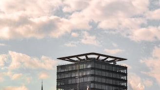 A modern glass-clad building with a distinctive flat roof structure set against a backdrop of a partly cloudy sky. Below the clouds, the building's simple geometric design contrasts with the organic, flowing shapes of the clouds.