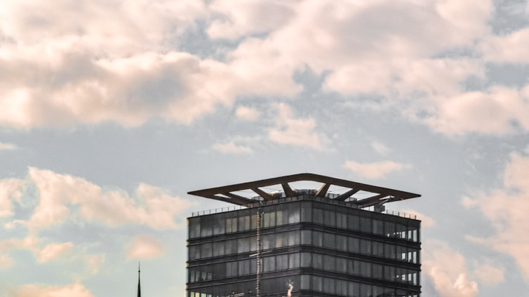 A modern glass-clad building with a distinctive flat roof structure set against a backdrop of a partly cloudy sky. Below the clouds, the building's simple geometric design contrasts with the organic, flowing shapes of the clouds.