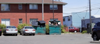 A parking lot with four parked cars in front of a brick building. The lot has two large green dumpsters that are overflowing with trash. Electrical wires and poles are visible, along with some overgrown vegetation around the dumpsters. Another building and a fence can be seen in the background.