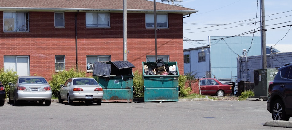 A parking lot with four parked cars in front of a brick building. The lot has two large green dumpsters that are overflowing with trash. Electrical wires and poles are visible, along with some overgrown vegetation around the dumpsters. Another building and a fence can be seen in the background.