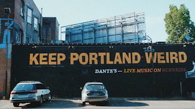 A large mural on the side of a building with the phrase 'KEEP PORTLAND WEIRD' in bold yellow letters. Below, in smaller font, it reads 'DANTE'S - LIVE MUSIC ON BURNSIDE'. Two parked cars are visible in the foreground, and there's an urban building structure in the background with a partially visible tree to the right.