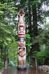 A tall, carved totem pole stands amidst a lush, green forest. The totem pole is decorated with colorful figures, each one stacked atop the other. Surrounding the totem pole are various trees and bushes, with a wooden pathway leading up to it. The area is serene and natural, with a combination of natural wooden elements and vibrant vegetation in the background.