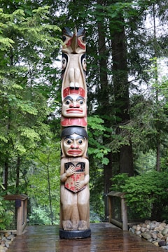 A tall, carved totem pole stands amidst a lush, green forest. The totem pole is decorated with colorful figures, each one stacked atop the other. Surrounding the totem pole are various trees and bushes, with a wooden pathway leading up to it. The area is serene and natural, with a combination of natural wooden elements and vibrant vegetation in the background.