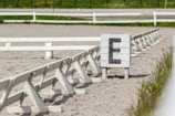 A sandy arena fenced with white wooden barriers, featuring a marker with the letter 'E' painted in black. The background shows a grassy field with some sparse vegetation.
