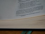Open book with printed text visible on the right side of the page. The text appears to be from a religious or philosophical context, and the book is set on a wooden surface with a green ribbon bookmark.