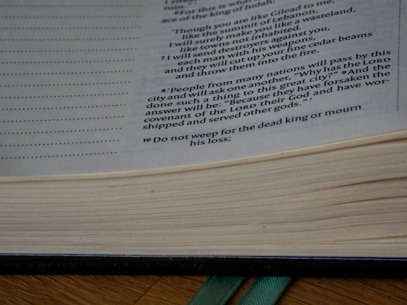 Open book with printed text visible on the right side of the page. The text appears to be from a religious or philosophical context, and the book is set on a wooden surface with a green ribbon bookmark.