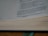 Open book with printed text visible on the right side of the page. The text appears to be from a religious or philosophical context, and the book is set on a wooden surface with a green ribbon bookmark.