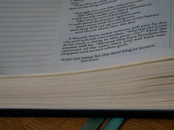 Open book with printed text visible on the right side of the page. The text appears to be from a religious or philosophical context, and the book is set on a wooden surface with a green ribbon bookmark.