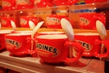 Red ceramic cups featuring labels resembling sardine cans are arranged neatly in rows. Each cup includes a white spoon tucked into its handle, creating a uniform and quirky display.