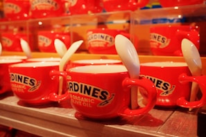 Red ceramic cups featuring labels resembling sardine cans are arranged neatly in rows. Each cup includes a white spoon tucked into its handle, creating a uniform and quirky display.