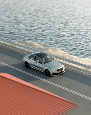 A silver sedan is captured driving along a coastal road with a view of the expansive ocean to the right. The car is moving on a smooth, dual-lane roadway, and a red-tiled roof is partially visible in the foreground.