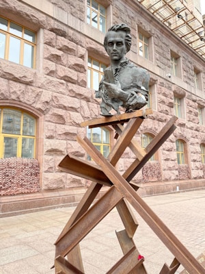 A modern sculpture is positioned in front of a building with textured stonework and yellow-trimmed windows. The sculpture consists of a metallic bust of a person with a thoughtful expression and folded arms, perched atop a series of intersecting metal beams.