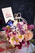 A vibrant bouquet of mixed flowers including roses, daisies, and chrysanthemums arranged in a decorative holder. The flowers vary in color from pink, peach, white, and purple. A card with the words 'Mamá Susana' and a heart symbol is inserted into the arrangement. The background is blurred, focusing on the floral display.