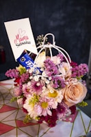 A vibrant bouquet of mixed flowers including roses, daisies, and chrysanthemums arranged in a decorative holder. The flowers vary in color from pink, peach, white, and purple. A card with the words 'Mamá Susana' and a heart symbol is inserted into the arrangement. The background is blurred, focusing on the floral display.