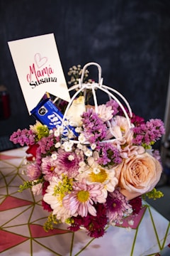 A vibrant bouquet of mixed flowers including roses, daisies, and chrysanthemums arranged in a decorative holder. The flowers vary in color from pink, peach, white, and purple. A card with the words 'Mamá Susana' and a heart symbol is inserted into the arrangement. The background is blurred, focusing on the floral display.