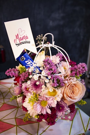 A vibrant bouquet of mixed flowers including roses, daisies, and chrysanthemums arranged in a decorative holder. The flowers vary in color from pink, peach, white, and purple. A card with the words 'Mamá Susana' and a heart symbol is inserted into the arrangement. The background is blurred, focusing on the floral display.