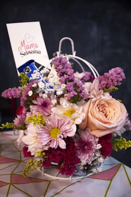 A beautifully arranged floral bouquet featuring a variety of flowers in shades of pink, peach, white, and purple. The bouquet includes roses, daisies, and other blossoms with mixed green foliage. A small card is inserted into the bouquet with a handwritten message and a heart design.