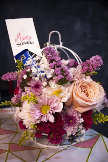 A beautifully arranged floral bouquet featuring a variety of flowers in shades of pink, peach, white, and purple. The bouquet includes roses, daisies, and other blossoms with mixed green foliage. A small card is inserted into the bouquet with a handwritten message and a heart design.