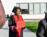 A person wearing a graduation cap and gown holds a diploma while standing outside. The gown is black with red accents. In the background, there are a few people sitting on a bench and a modern building with large windows.