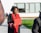 A person wearing a graduation cap and gown holds a diploma while standing outside. The gown is black with red accents. In the background, there are a few people sitting on a bench and a modern building with large windows.