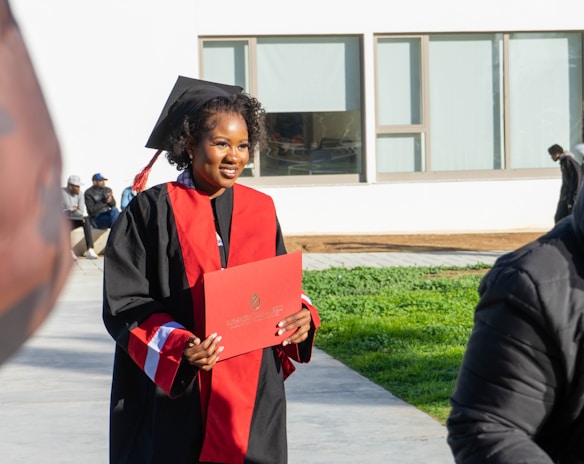 A person wearing a graduation cap and gown holds a diploma while standing outside. The gown is black with red accents. In the background, there are a few people sitting on a bench and a modern building with large windows.