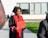 A person wearing a graduation cap and gown holds a diploma while standing outside. The gown is black with red accents. In the background, there are a few people sitting on a bench and a modern building with large windows.