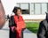 A person wearing a graduation cap and gown holds a diploma while standing outside. The gown is black with red accents. In the background, there are a few people sitting on a bench and a modern building with large windows.