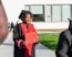 A person wearing a graduation cap and gown holds a diploma while standing outside. The gown is black with red accents. In the background, there are a few people sitting on a bench and a modern building with large windows.