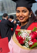 A young woman is wearing a graduation cap and gown, holding a bouquet of red roses and white flowers. She is smiling and standing outdoors among other graduates. The background features a modern glass building and several people dressed in similar attire.
