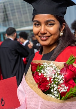 A young woman is wearing a graduation cap and gown, holding a bouquet of red roses and white flowers. She is smiling and standing outdoors among other graduates. The background features a modern glass building and several people dressed in similar attire.