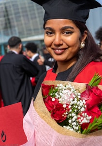 A young woman is wearing a graduation cap and gown, holding a bouquet of red roses and white flowers. She is smiling and standing outdoors among other graduates. The background features a modern glass building and several people dressed in similar attire.
